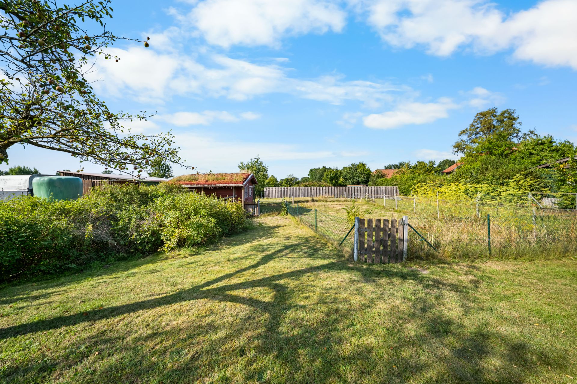 Großer Garten mit Wiese, Sträuchern, Holzzaun und kleiner Hütte im Hintergrund.