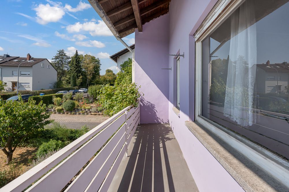 Balkon mit lila Wand, Blick auf Garten und Nachbarhäuser, teils bewölkter Himmel.
