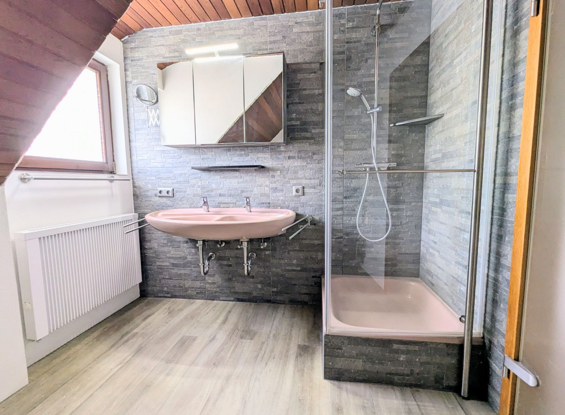 Helles Bad mit rosafarbenem Doppelwaschbecken und Dusche, Spiegel und Fenster.