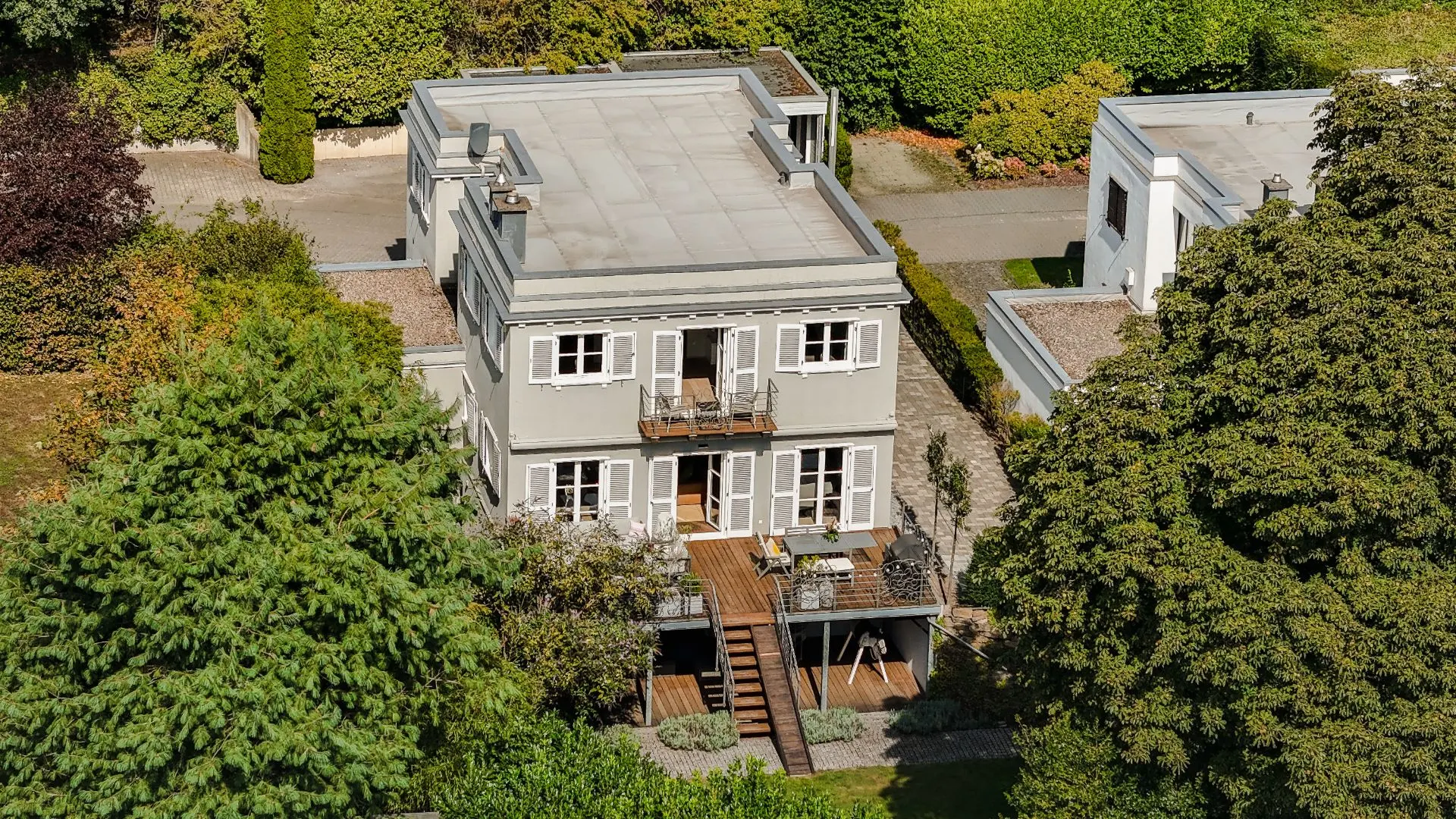 Modernes zweistöckiges Wohnhaus mit Flachdach, Balkon und Terrasse, umgeben von Bäumen und Garten.