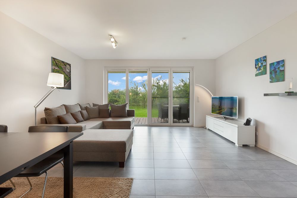 Helles Wohnzimmer mit Ecksofa, Fernseher, Esstisch und großen Fenstern zum Garten.