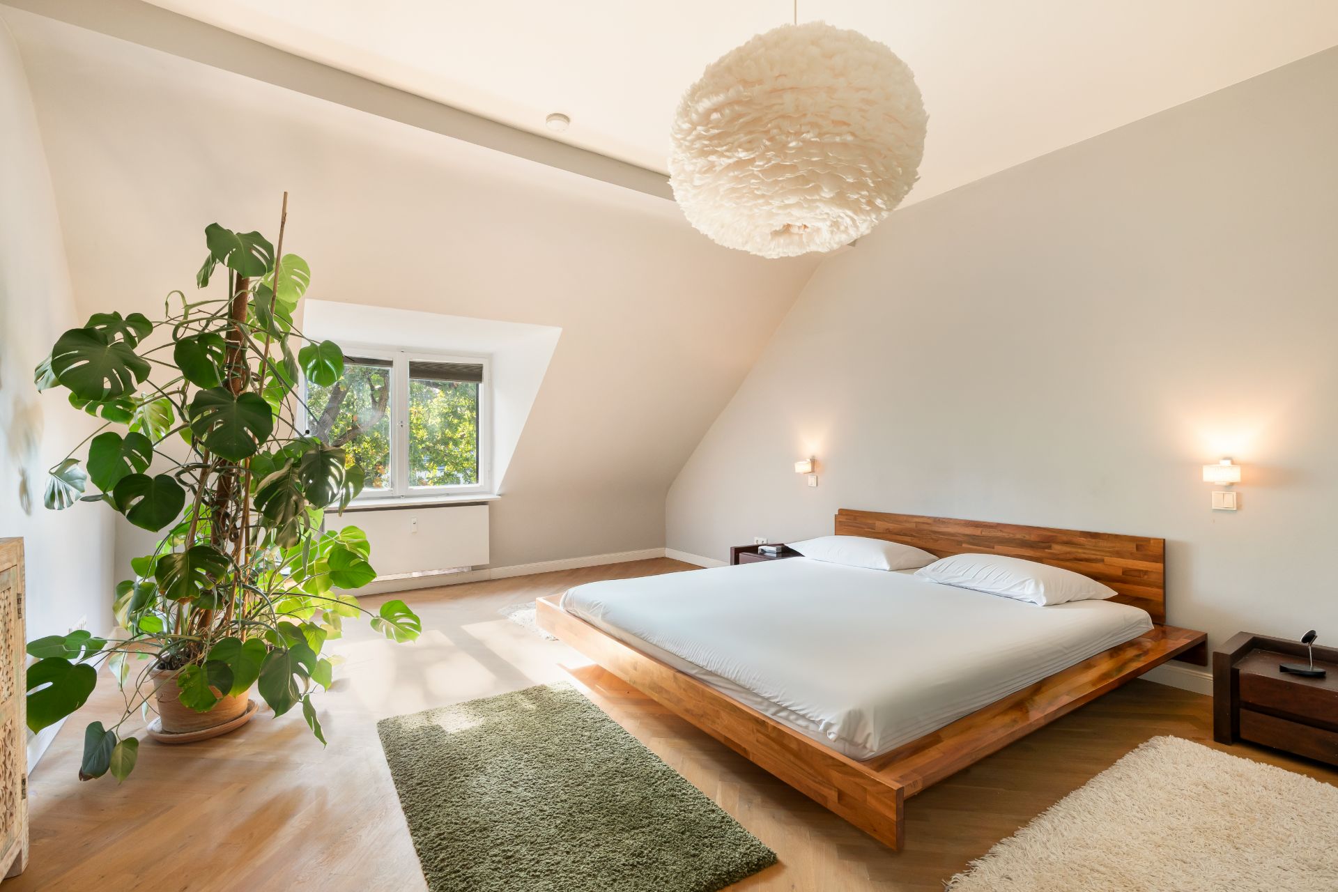 Helles Schlafzimmer mit Holzbett, großem Fenster, zwei Teppichen und einer großen Zimmerpflanze.