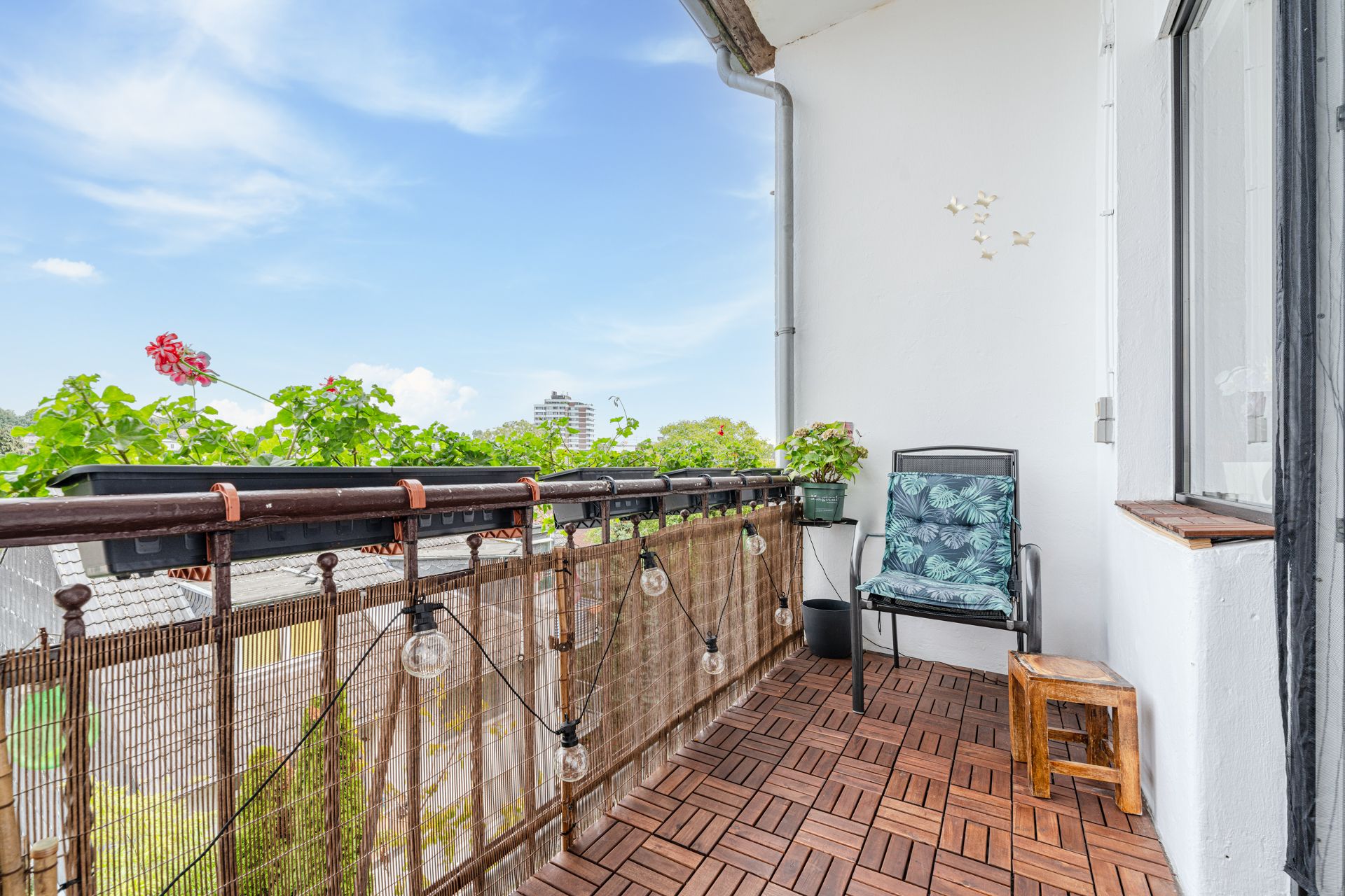 Balkon mit Holzfliesen, einem Stuhl, Blumen, Lichterkette und Ausblick ins Grüne.