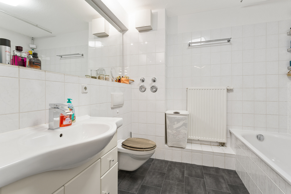 Modernes Badezimmer mit Waschbecken, WC, Badewanne und Wäschekorb, weiße Fliesen.