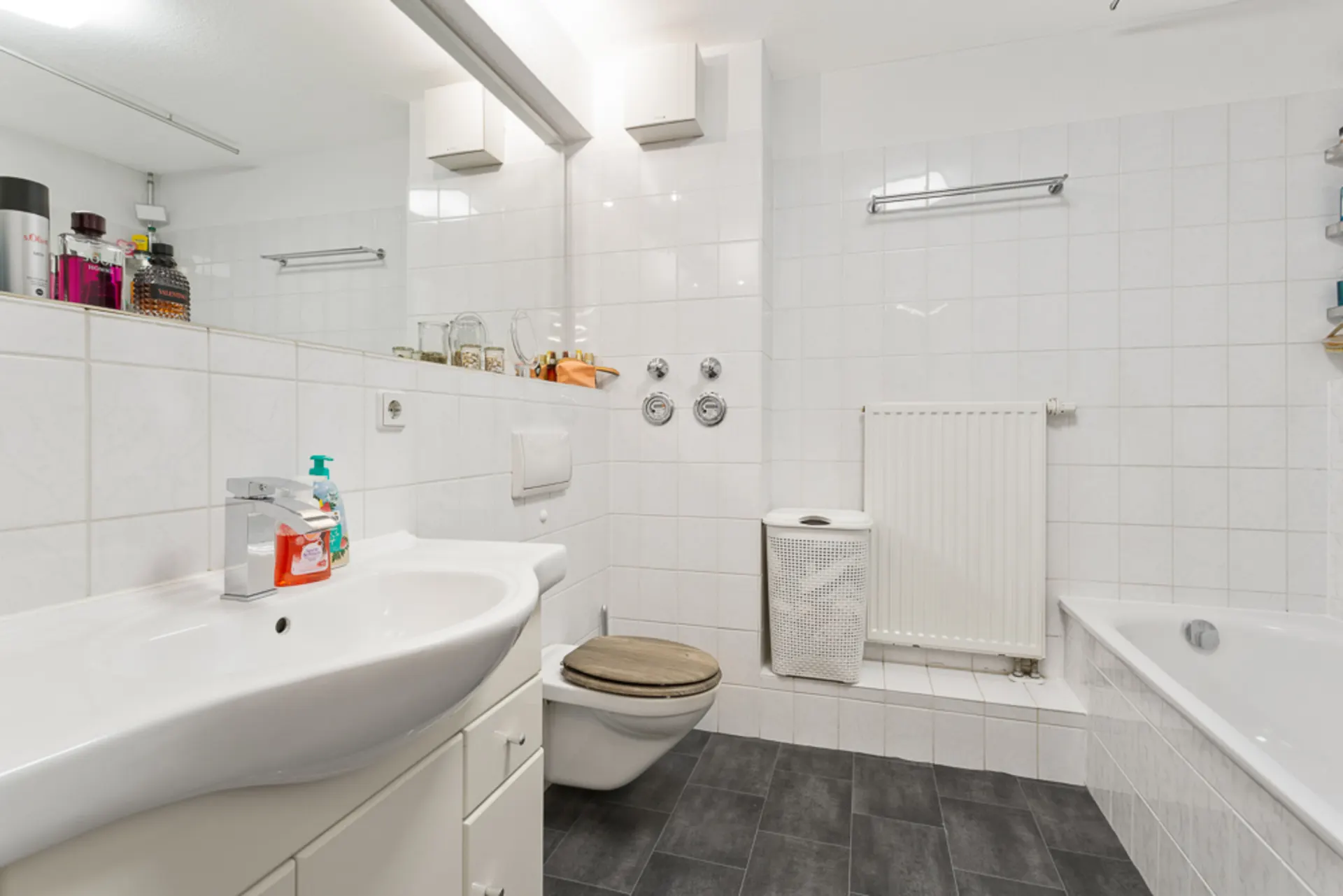 Modernes Badezimmer mit Waschbecken, WC, Badewanne und Wäschekorb, weiße Fliesen.