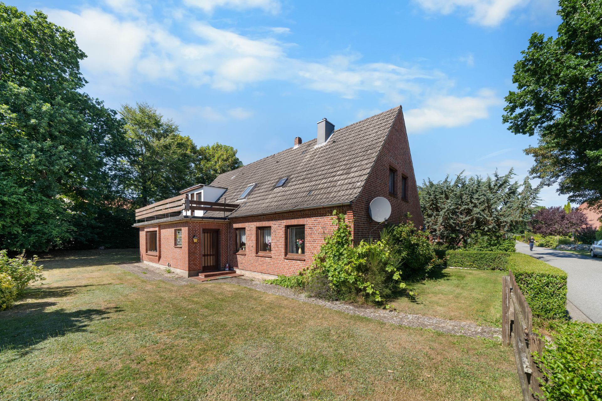 Backsteinwohnhaus mit Satteldach, großer Rasenfläche und umliegenden Bäumen an ruhiger Straße.