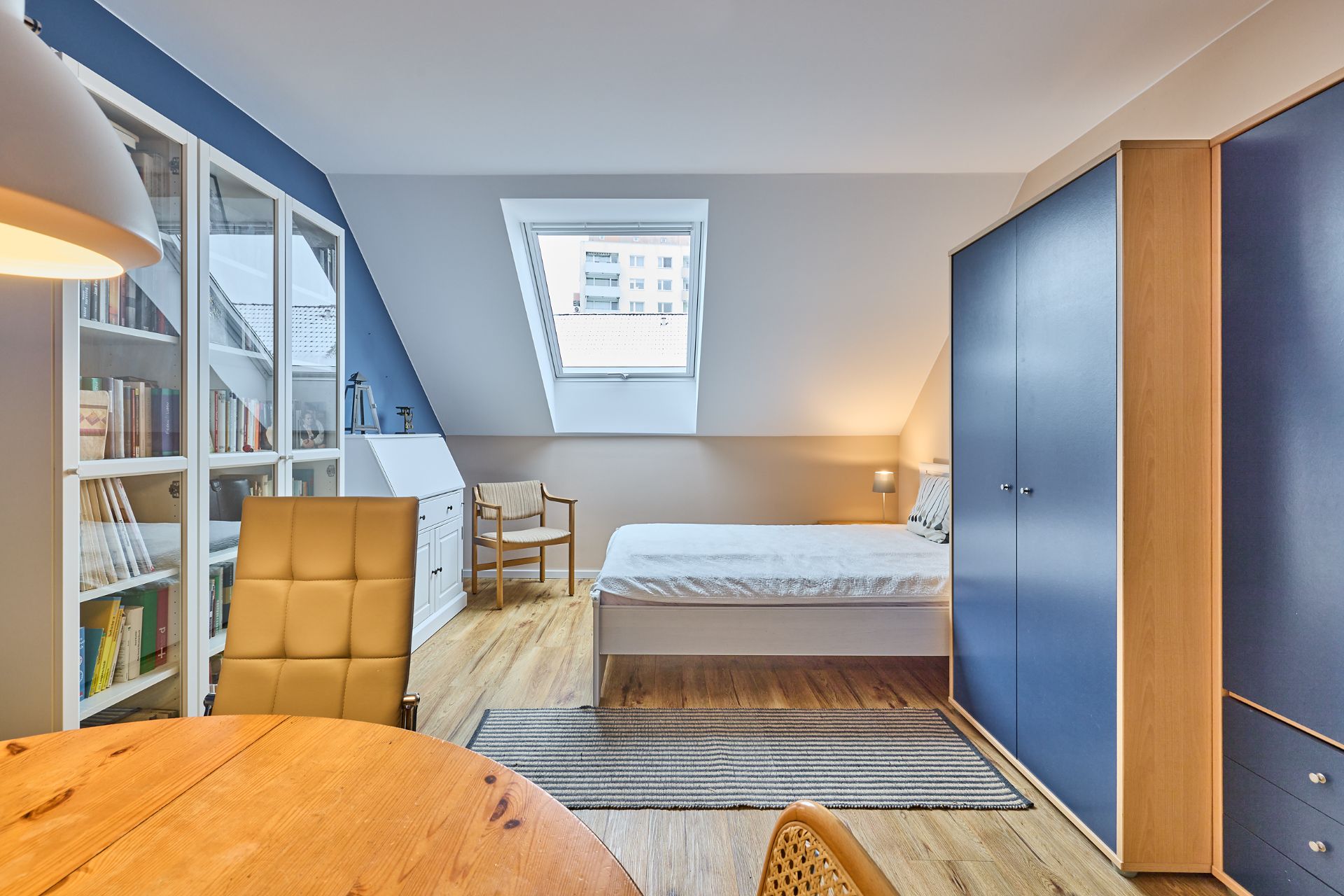 Helles Schlafzimmer mit Bett, Schrank, Bücherregal, Tisch und Dachfenster.