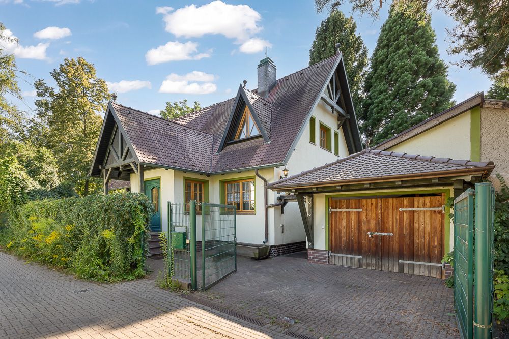 Einfamilienhaus mit Satteldach, Holzgarage und grünem Zaun, umgeben von Bäumen.