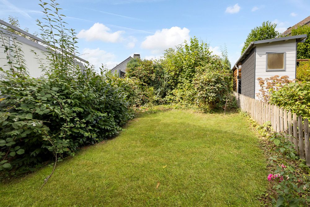 Grüner Garten mit Rasenfläche, Büschen, kleinem Holzzaun und Gartenhaus bei Tageslicht.