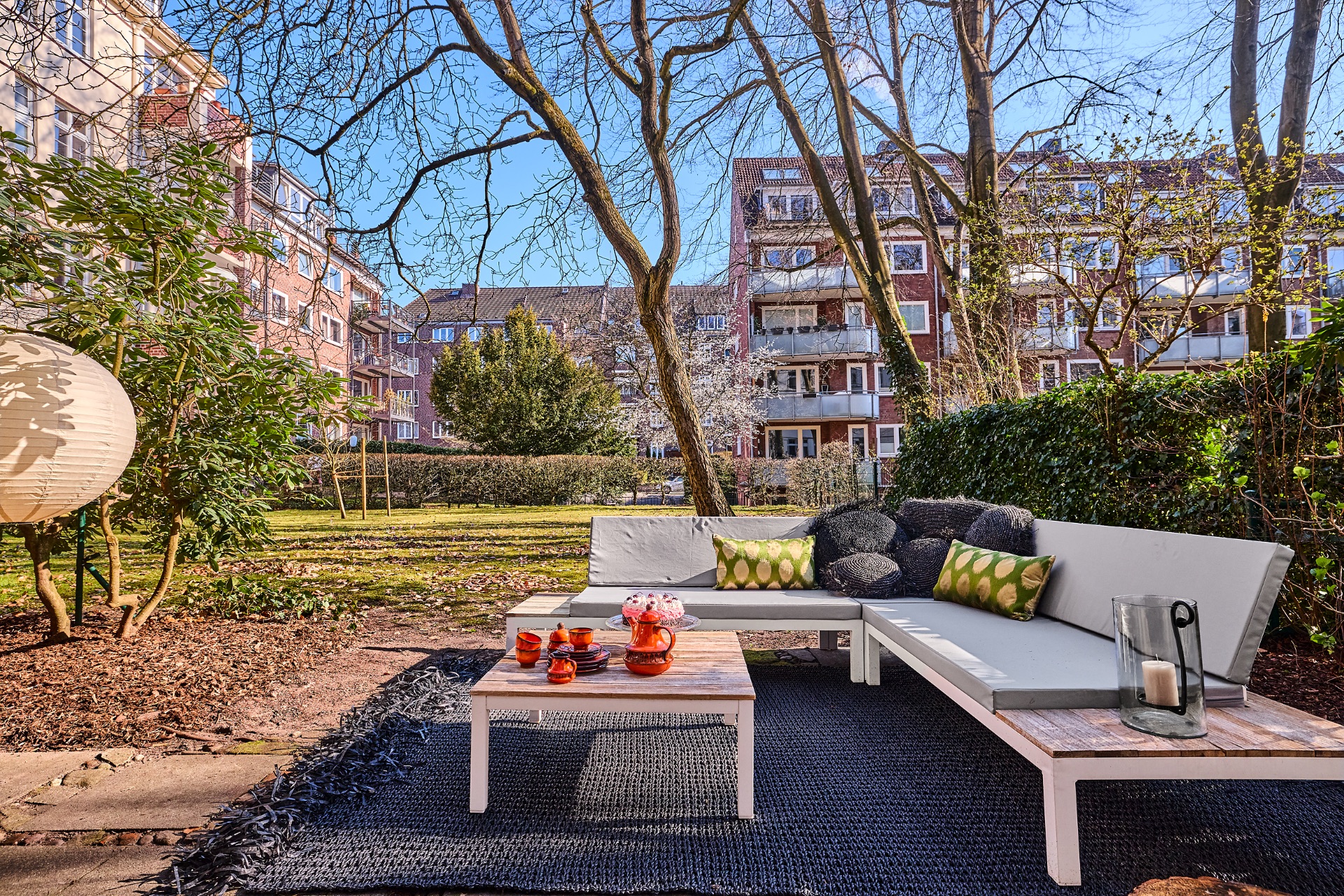 Garten mit Sitzgruppe, kleinem Tisch und Kissen, im Hintergrund Wohngebäude und Bäume.