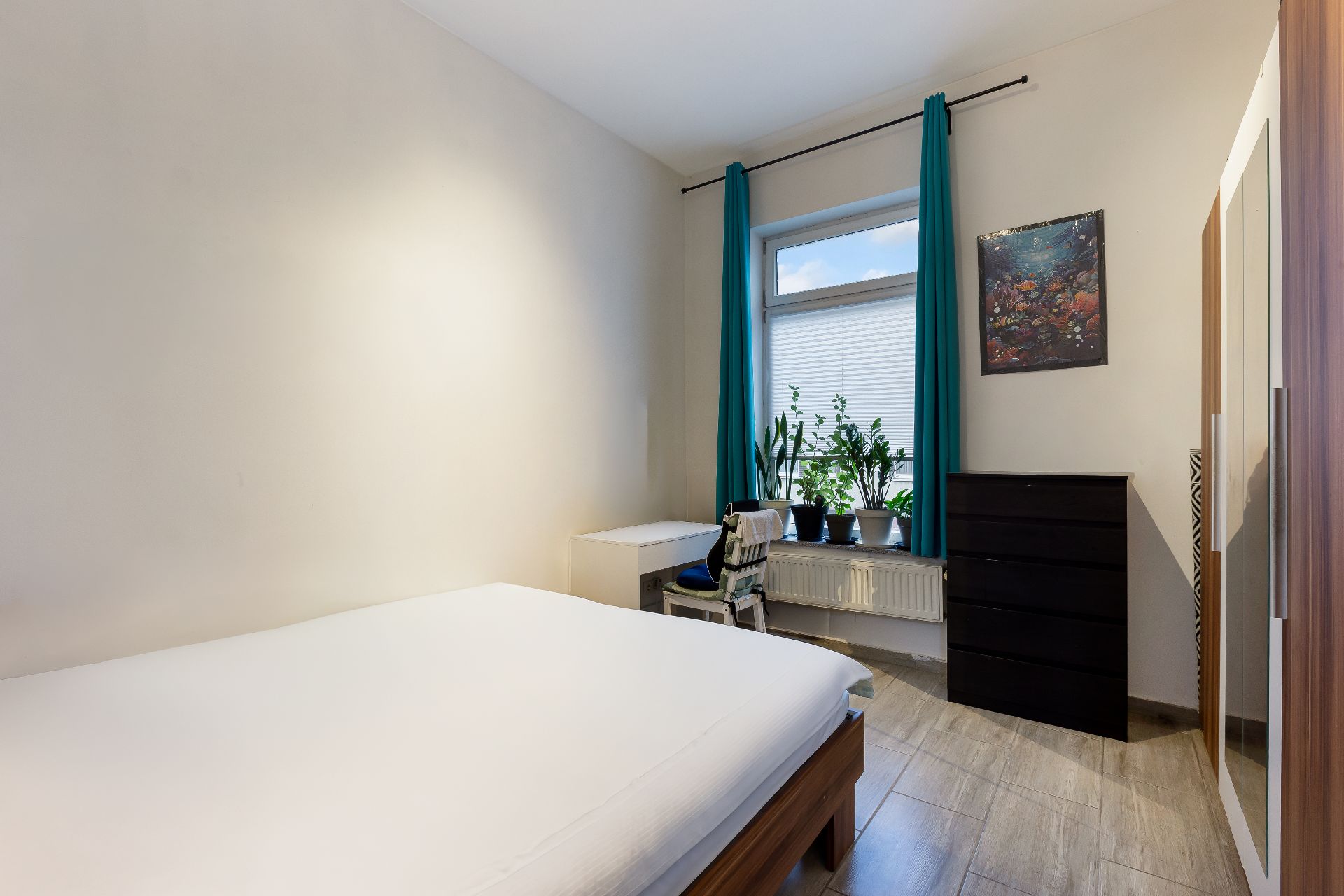 Helles Schlafzimmer mit Bett, Schrank, Kommode, Schreibtisch und Fenster mit Pflanzen.