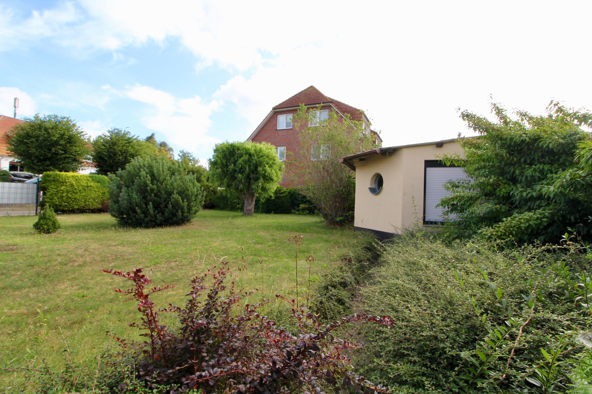 Garten mit Rasen, Büschen, Bäumen und zwei Häusern im Hintergrund.
