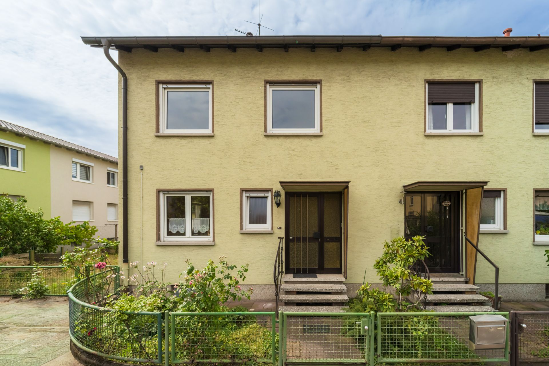 Vorderansicht eines zweistöckigen Reihenhauses mit kleinem Garten und grünem Zaun.