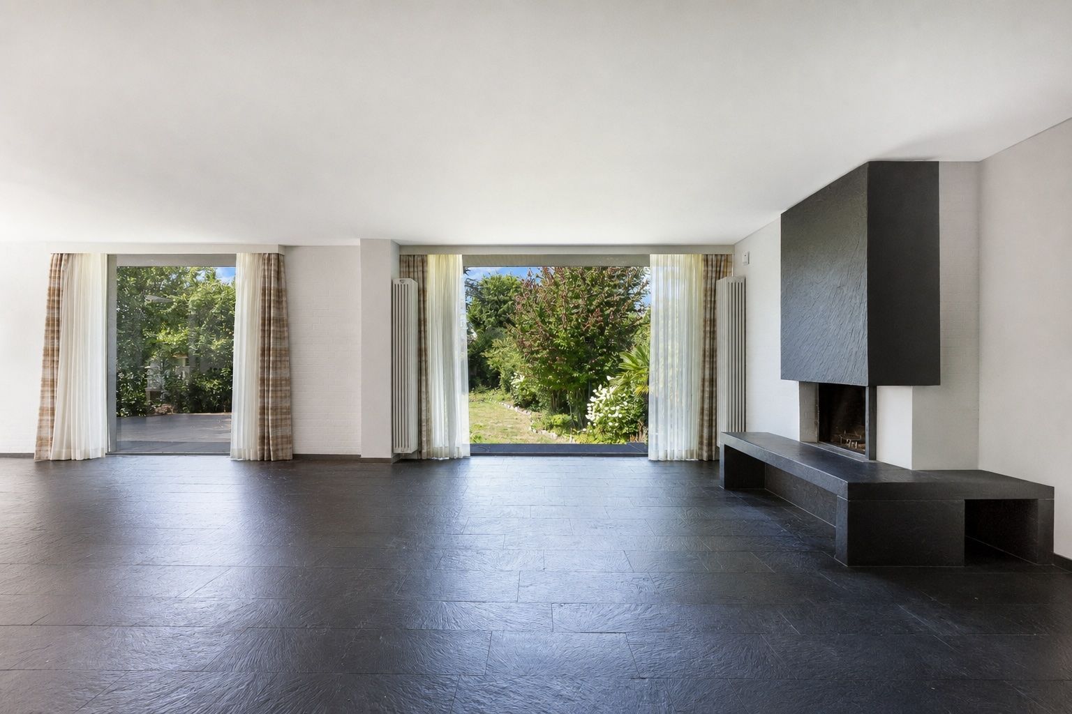 Helles Wohnzimmer mit großen Fenstern, Vorhängen und modernem Kamin. Blick auf Garten.