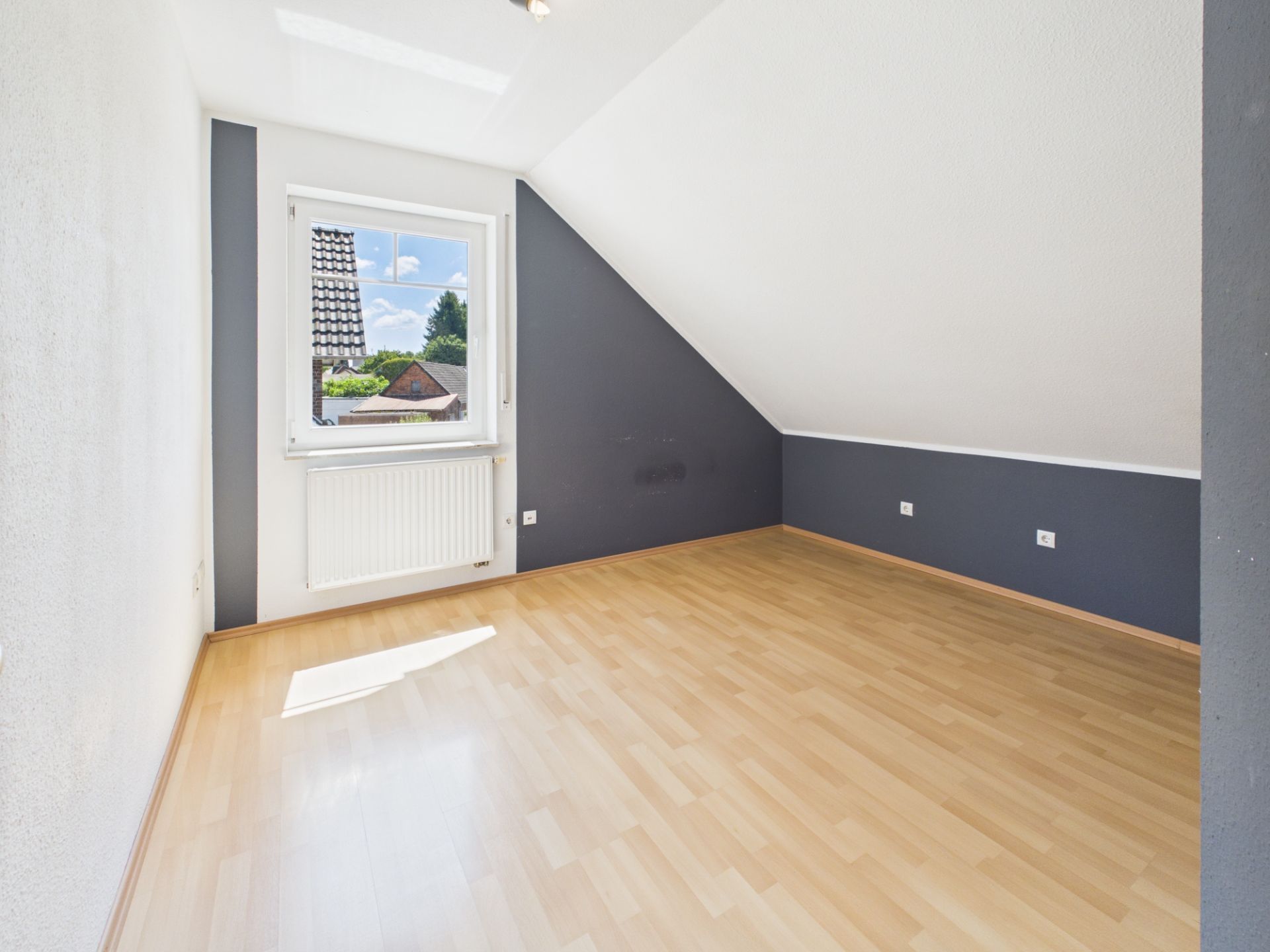 Leeres Zimmer mit schräger Decke, Holzfußboden und Fenster mit Blick nach draußen.