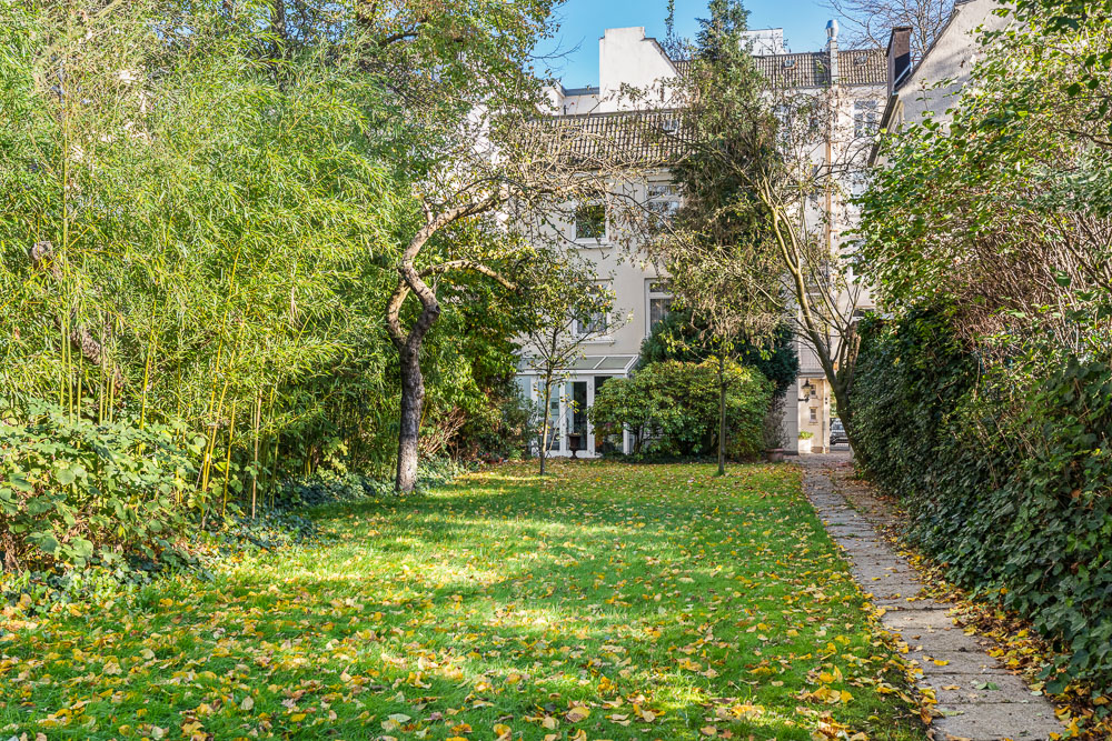 Grüner Garten mit Bäumen, Rasen und einem Weg entlang eines Wohnhauses.