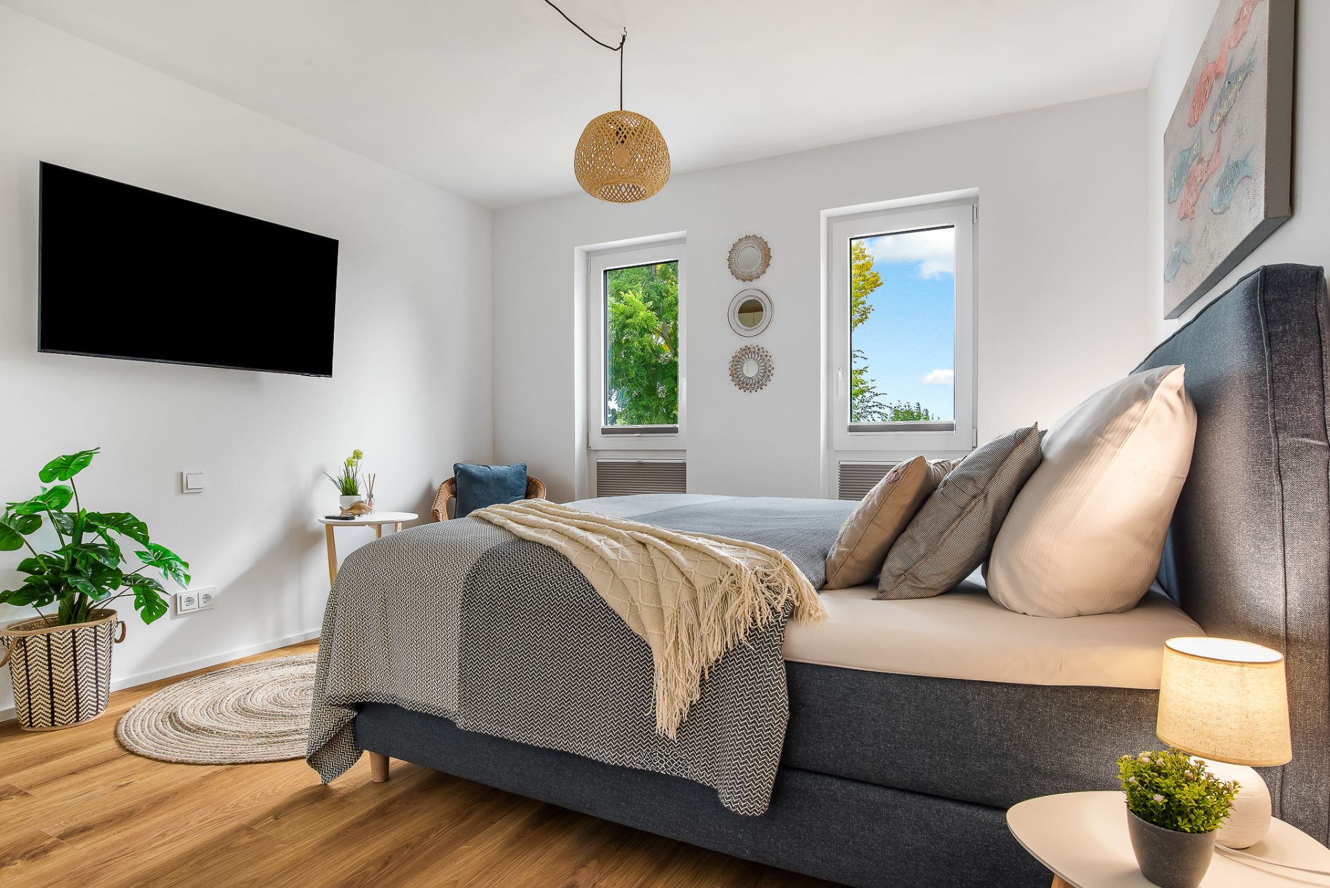 Modernes Schlafzimmer mit Doppelbett, Fernseher, Zimmerpflanzen und zwei Fenstern mit Blick ins Grüne.
