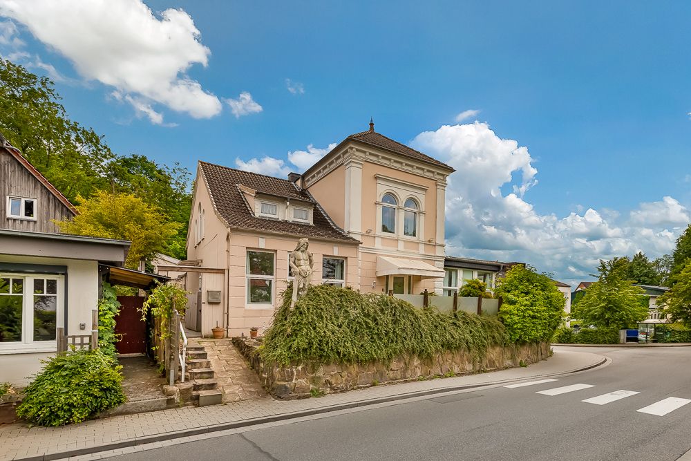 Zweistöckiges Wohnhaus mit Garten, Hecke und einer Statue vor dem Eingang an einer Straßenecke.