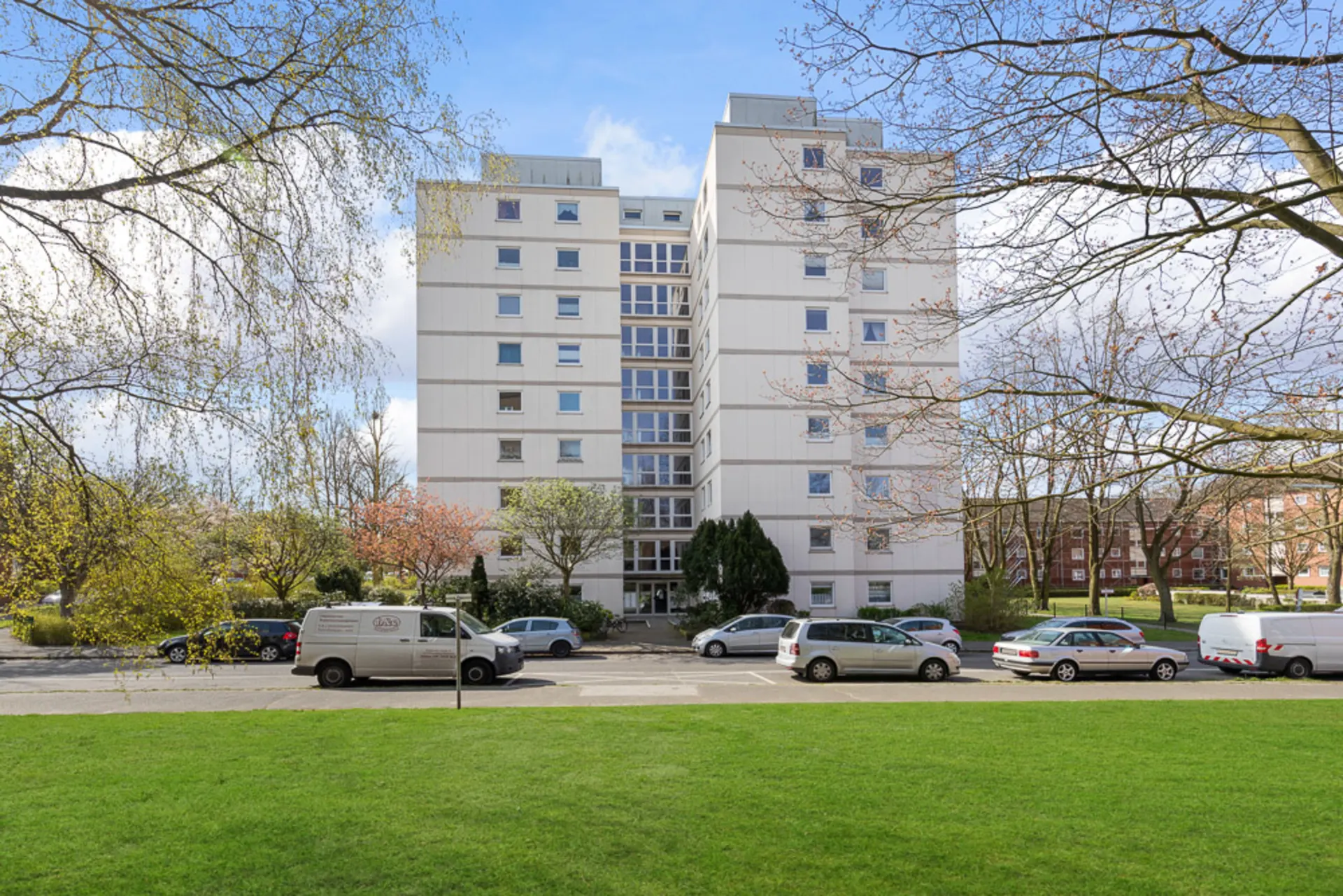 Mehrstöckiges Wohnhaus mit zahlreichen Fenstern, Autos parken davor, grüne Wiese im Vordergrund.