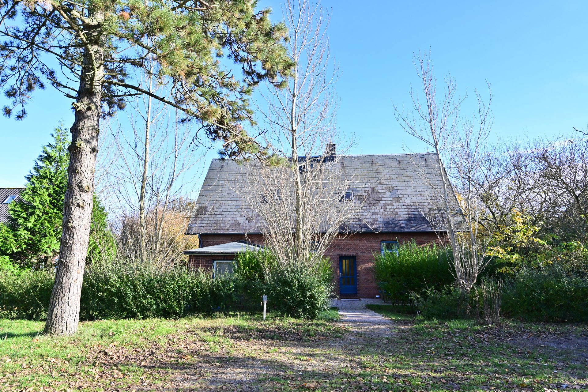 Backsteinhaus mit Satteldach, umgeben von Bäumen und Sträuchern, großer Garten im Vordergrund.