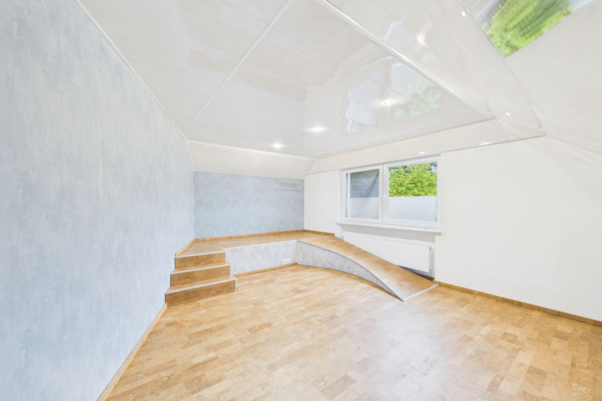 Leerer Raum mit Holzfußboden, Stufe, Podest und großem Fenster mit Blick ins Grüne.