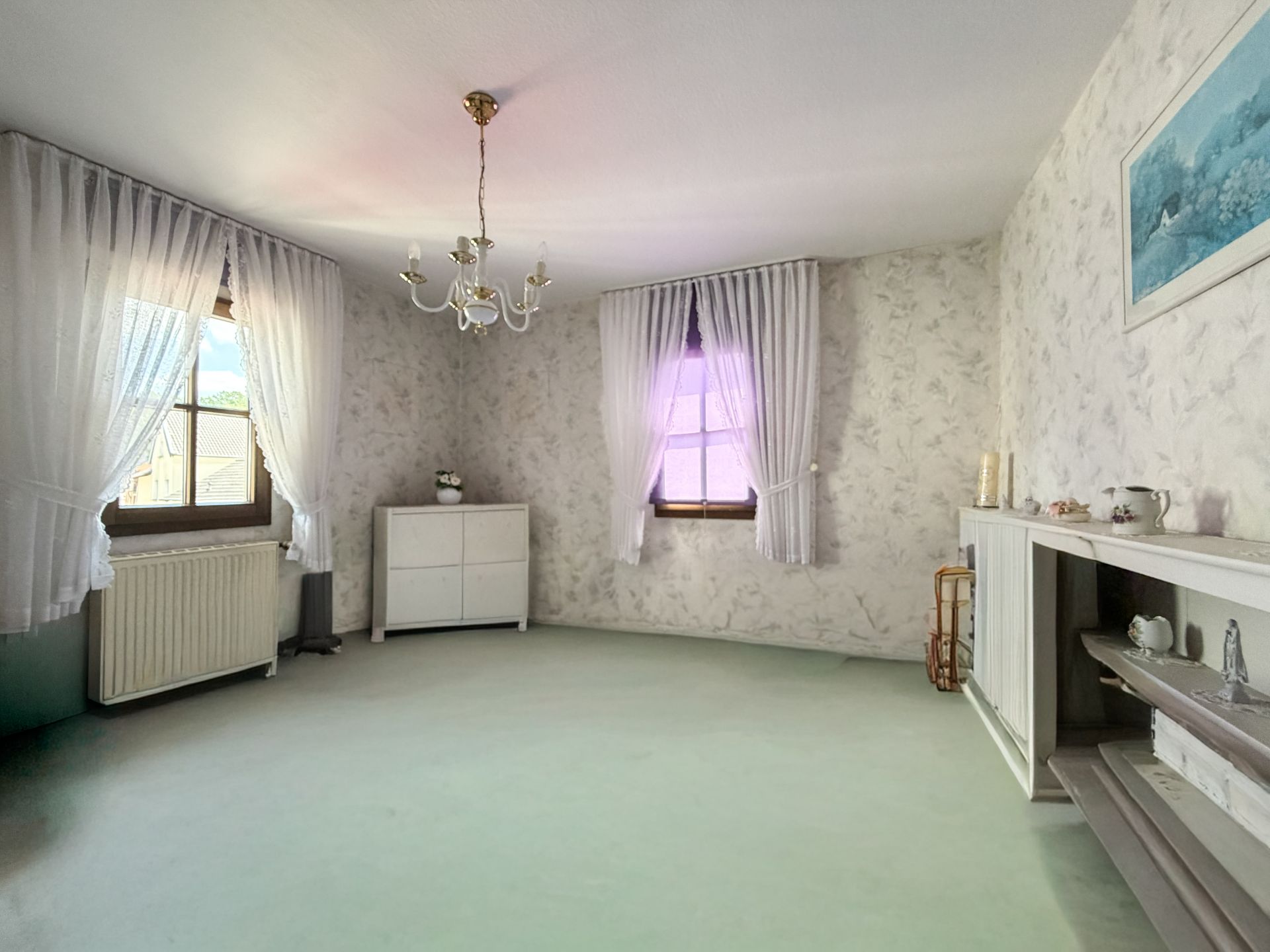 Helles Wohnzimmer mit zwei Fenstern, weißen Vorhängen, Kommode und Kaminregal.