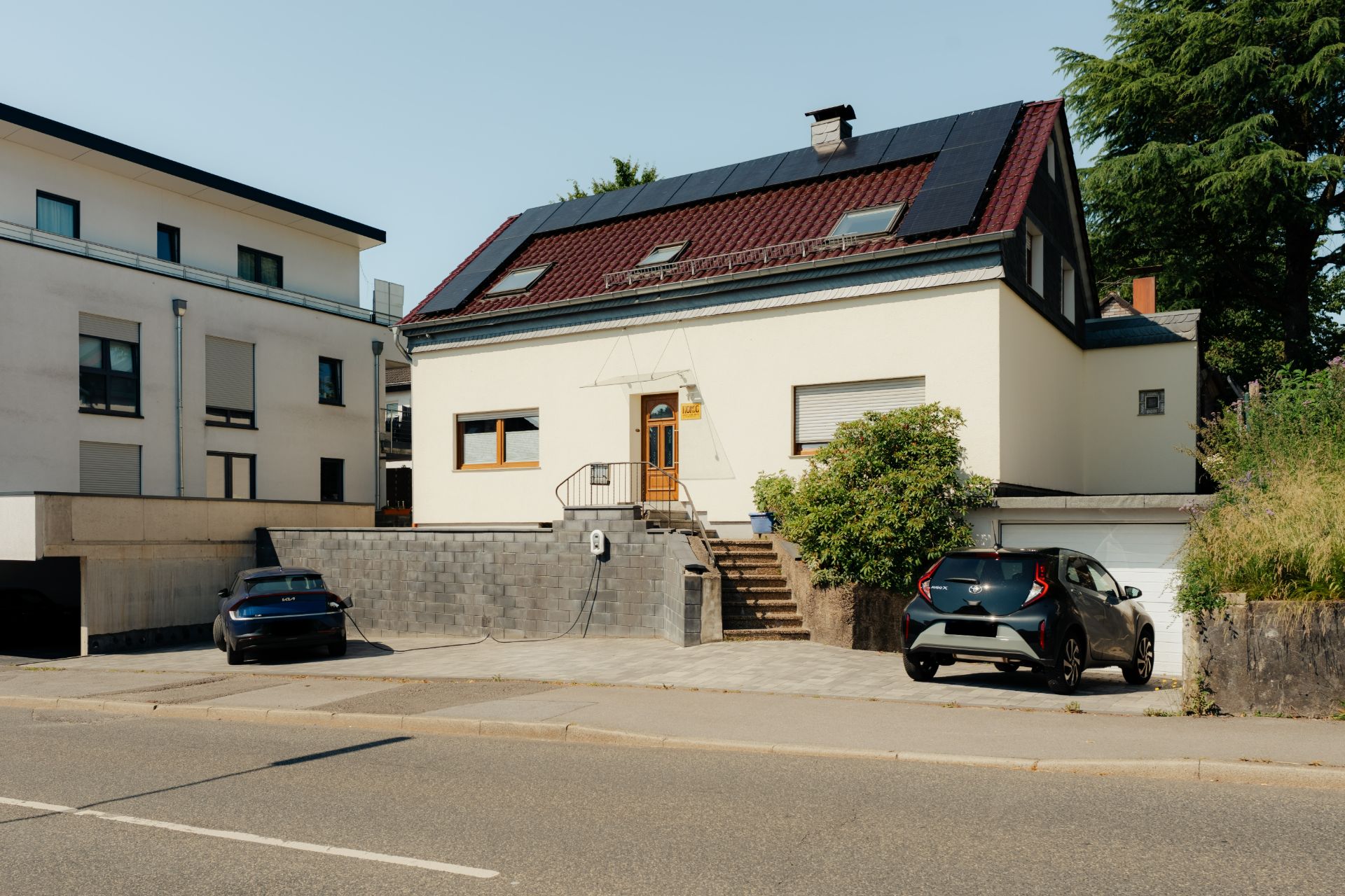 Zweistöckiges Wohnhaus mit rotem Dach, Solarpanels und zwei geparkten Autos vor dem Haus.