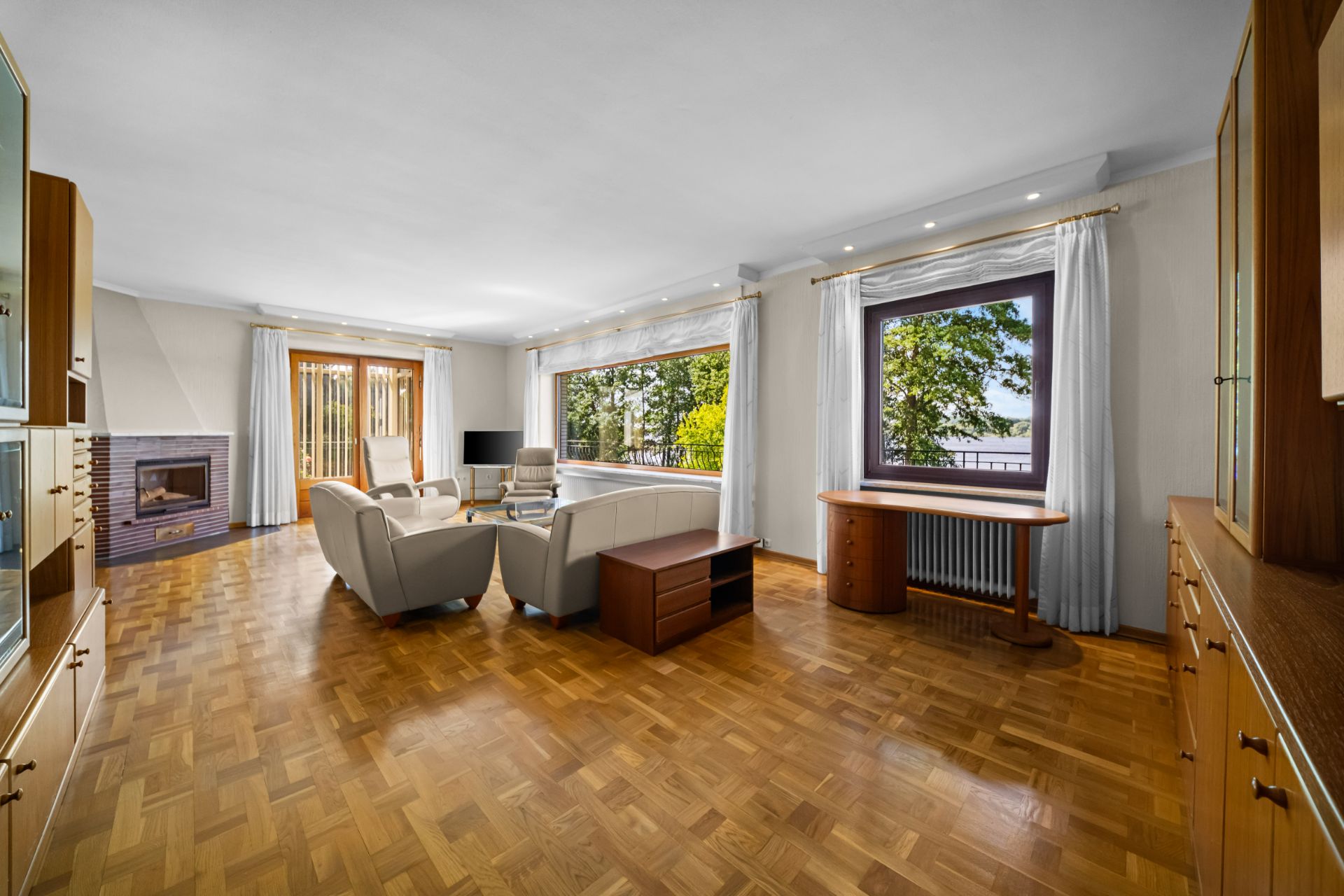 Helles Wohnzimmer mit Parkettboden, Sofas, Kamin, Schrankwänden und großen Fenstern mit Blick ins Grüne.
