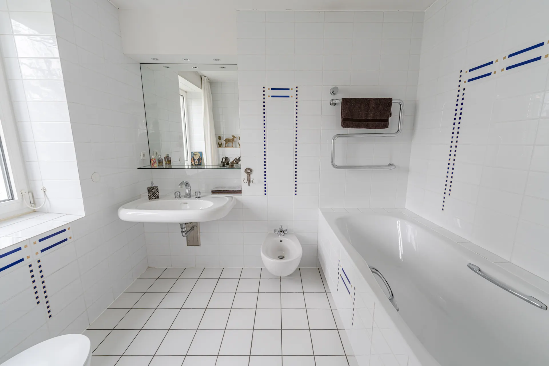 Helles Badezimmer mit Badewanne, Waschbecken, Bidet, großem Spiegel und weißen Fliesen.