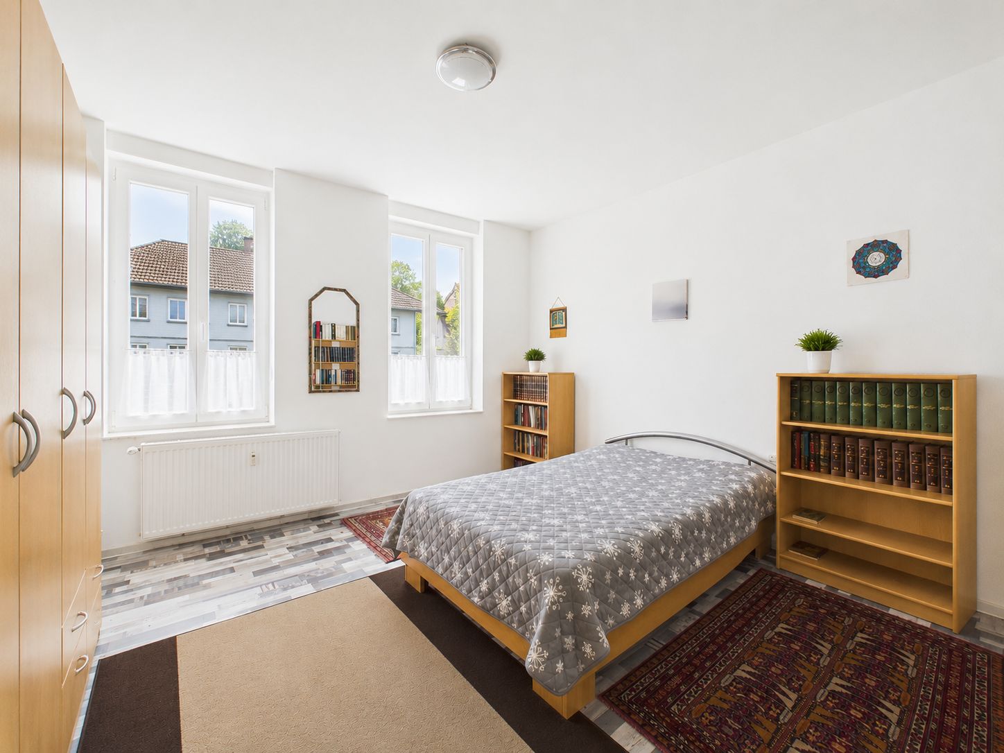 Helles Schlafzimmer mit Doppelbett, zwei Bücherregalen, Teppichen und großen Fenstern.