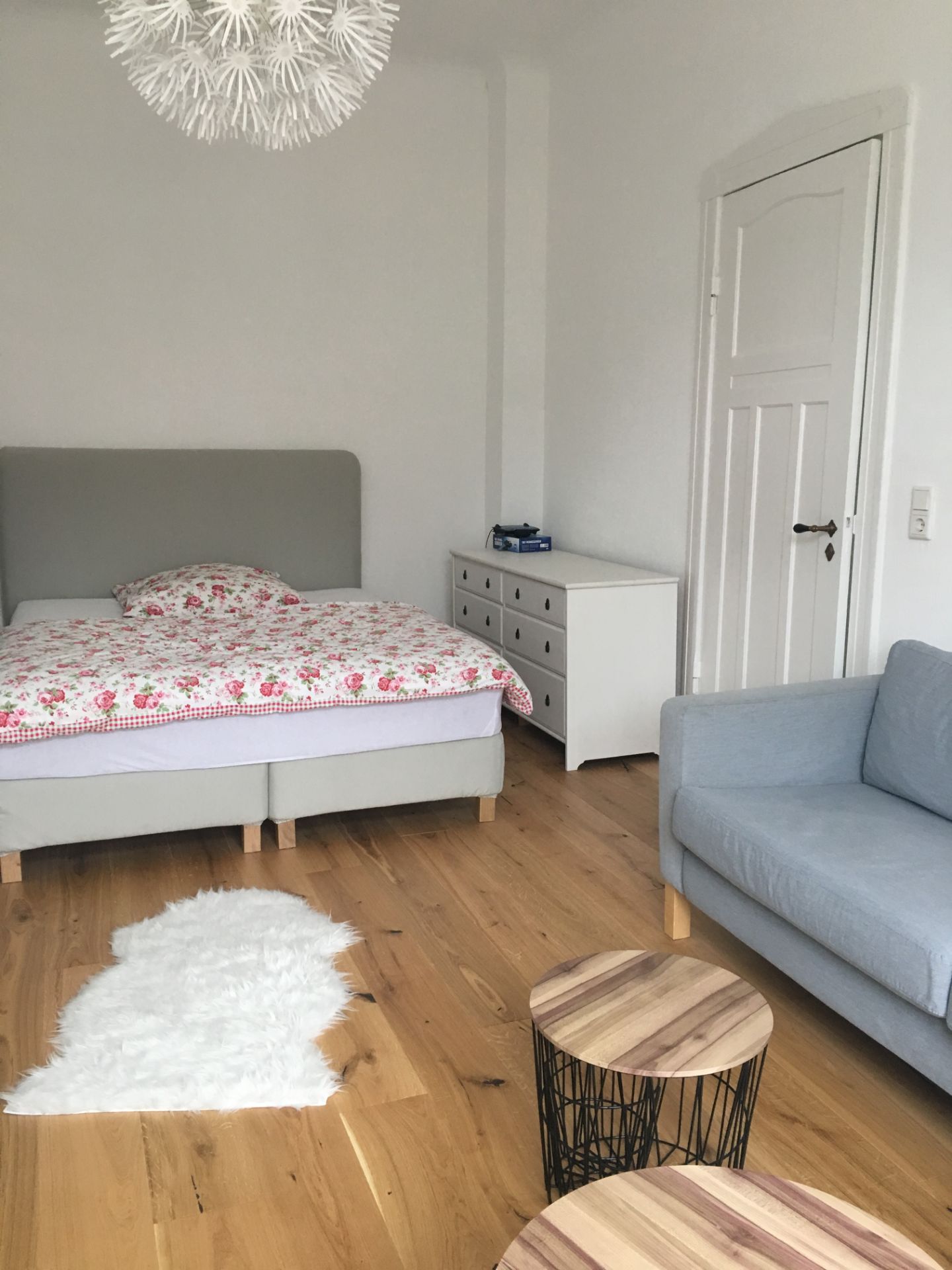 Helles Schlafzimmer mit Doppelbett, grauem Sofa, Kommode und zwei runden Holztischen auf Holzboden.