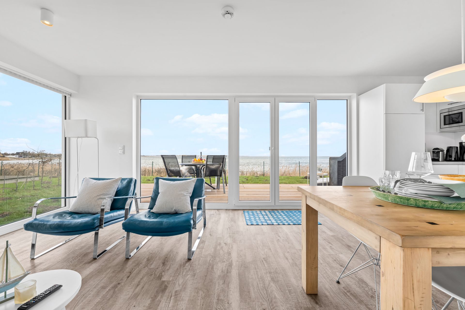 Helles Wohnzimmer mit zwei blauen Sesseln, Holztisch und Zugang zur Terrasse mit Meerblick.