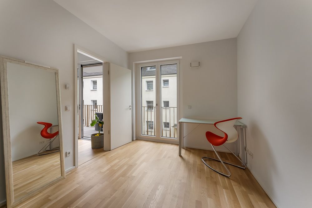 Helles Zimmer mit Holzboden, Spiegel, kleinem Schreibtisch und rotem Stuhl. Tür führt zu Balkon.