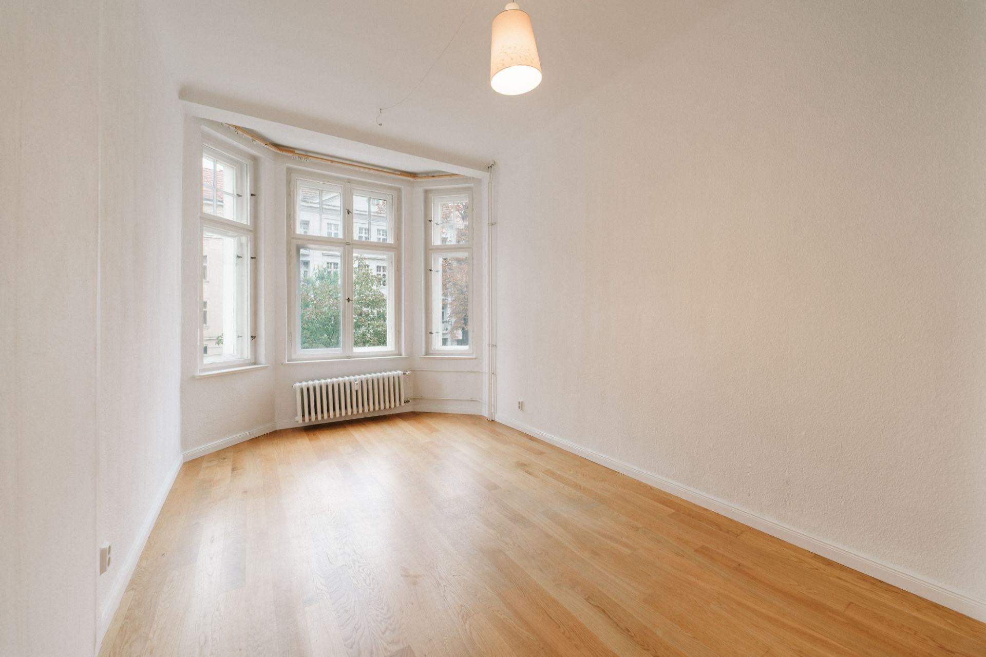 Leeres Zimmer mit Holzfußboden, großem Fenster, Heizung und Deckenlampe.