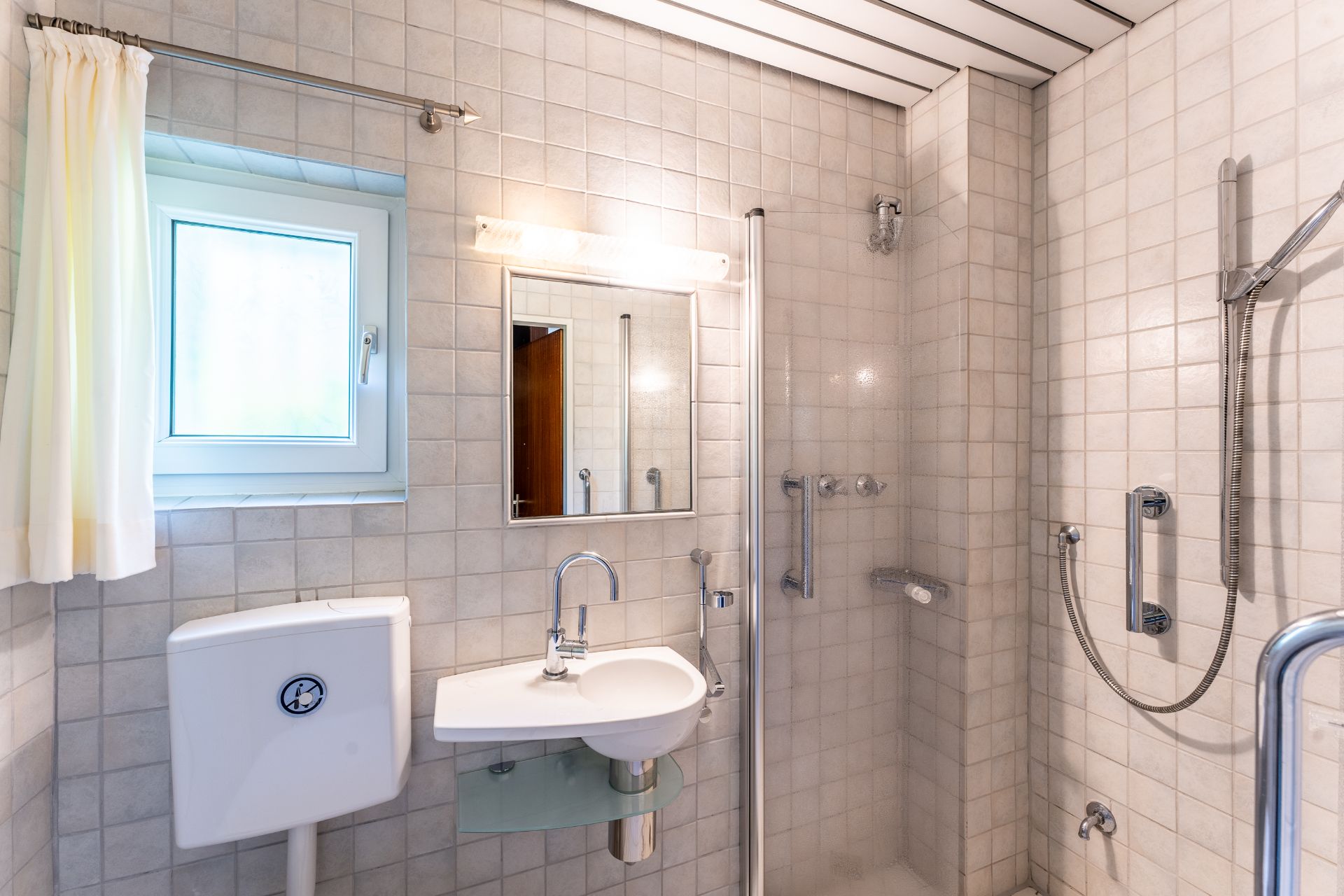 Helles Badezimmer mit Fenster, Spiegel, Waschbecken, Toilette und barrierefreier Dusche.