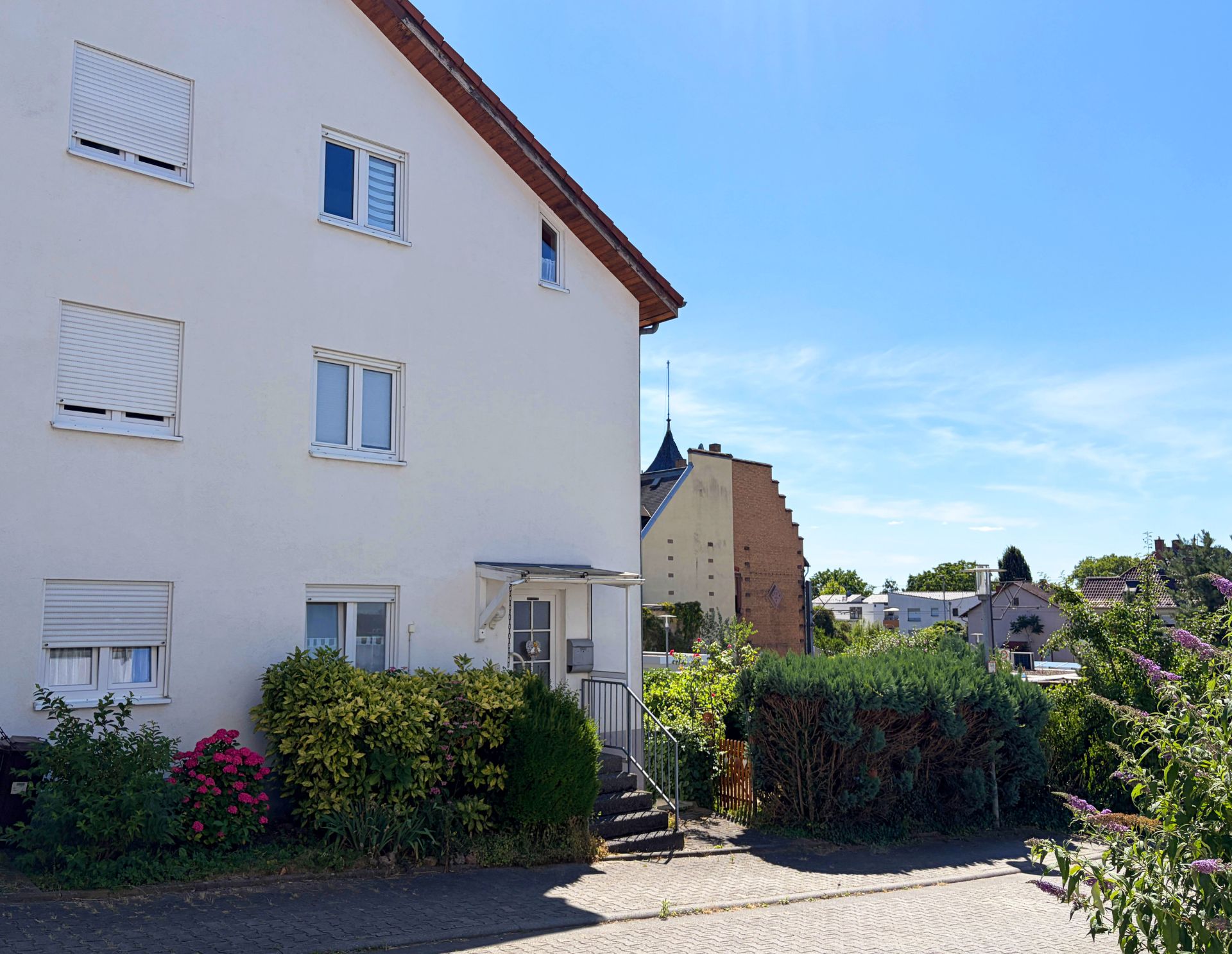 Helles Wohnhaus mit drei Stockwerken, Eingangstreppe und umliegendem Garten bei Sonnenschein.