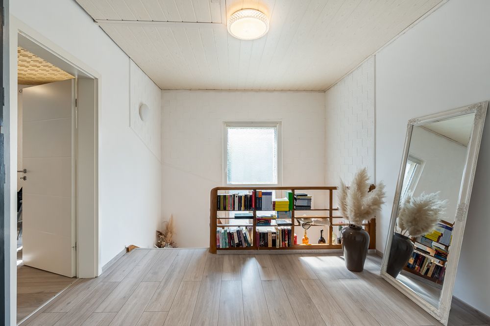 Heller Raum mit Regal voller Bücher, großem Spiegel und zwei Vasen mit Trockenblumen.