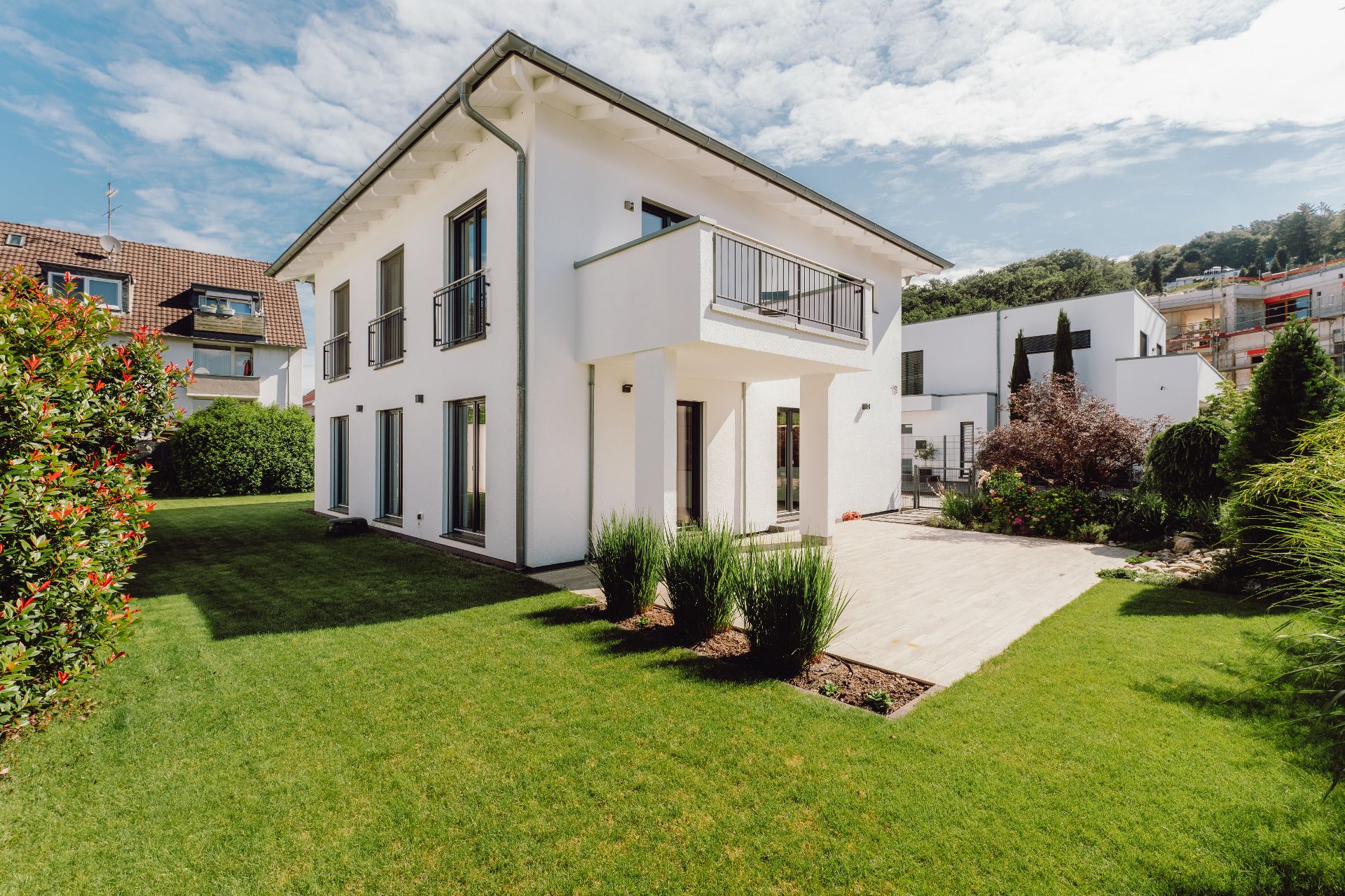 Modernes weißes Wohnhaus mit Balkon und gepflegtem Garten an einem sonnigen Tag.
