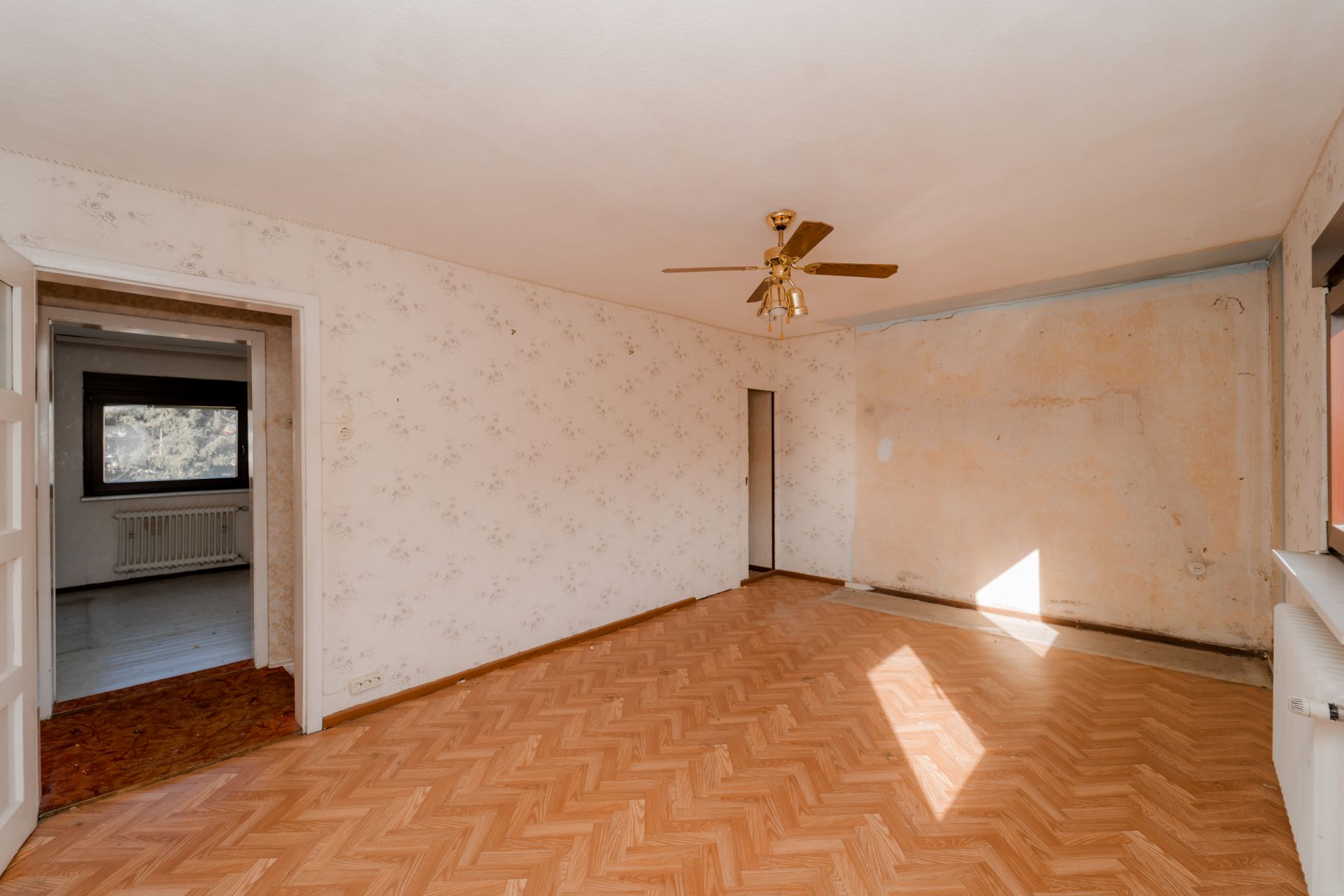 Leeres Zimmer mit Parkettboden, Deckenventilator und Tapete, Tür zum Nebenzimmer sichtbar.
