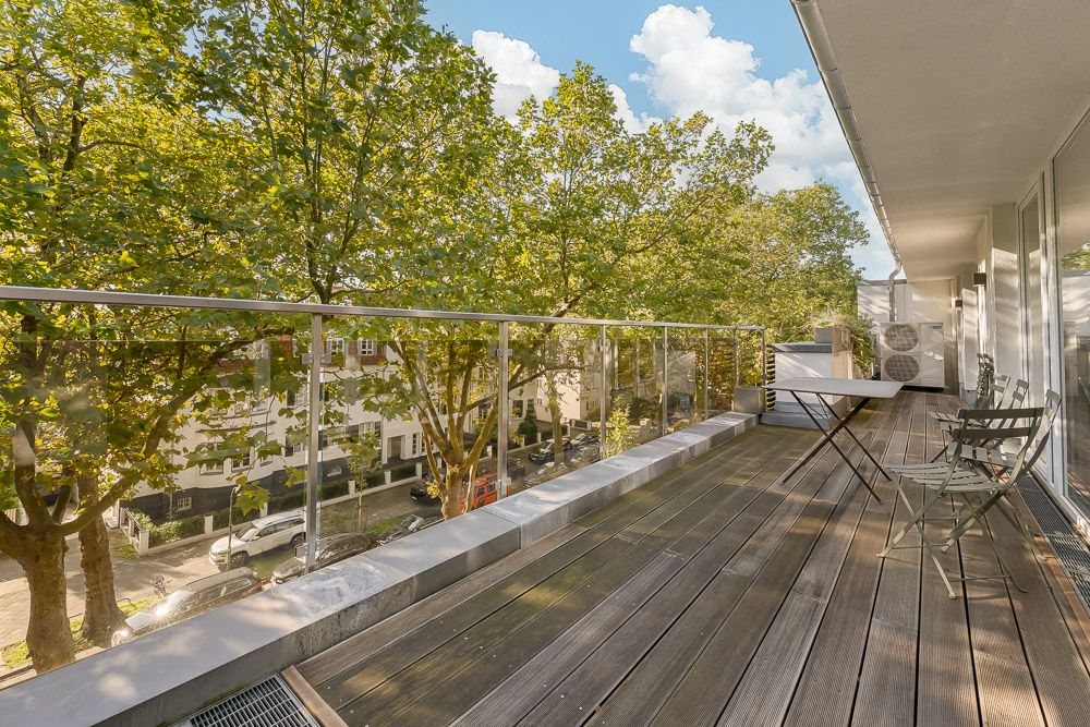 Großer Balkon mit Holzboden, Tisch und Stühlen, Ausblick auf Bäume und Wohnstraße.