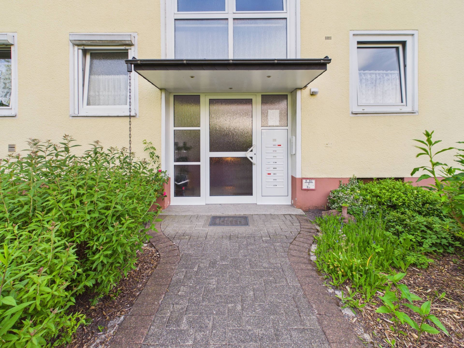 Eingang eines Mehrfamilienhauses mit Glas­tür, Briefkästen und umliegenden Pflanzen.