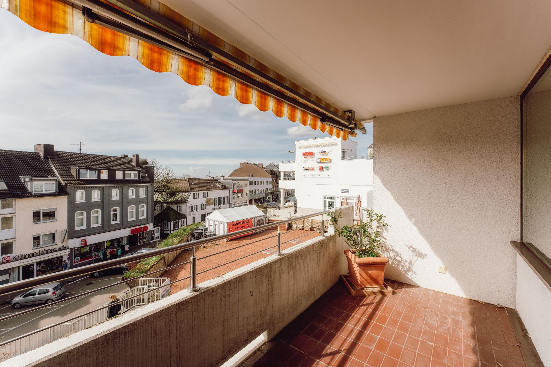Balkon mit Topfpflanze und Markise, Blick auf belebte Straße und Nachbargebäude.