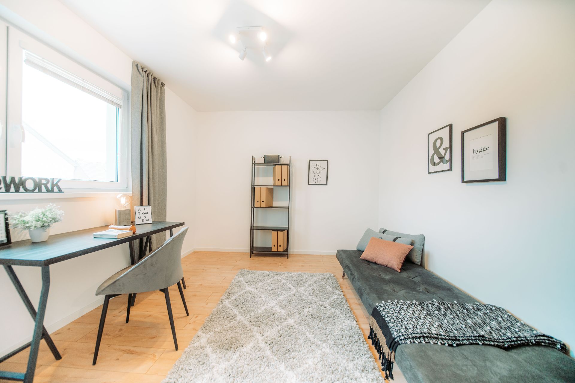 Helles Zimmer mit Schreibtisch, Regal, grauem Sofa und Teppich auf Holzfußboden.