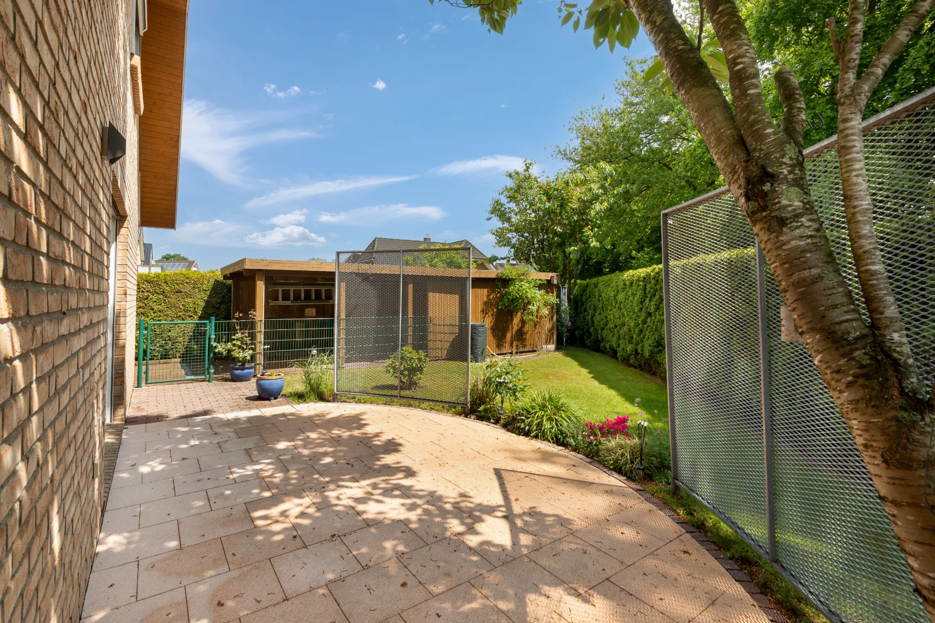 Gepflasterte Terrasse mit Zaun, Baum, Gartenhaus und grünem Rasen im Hintergrund.