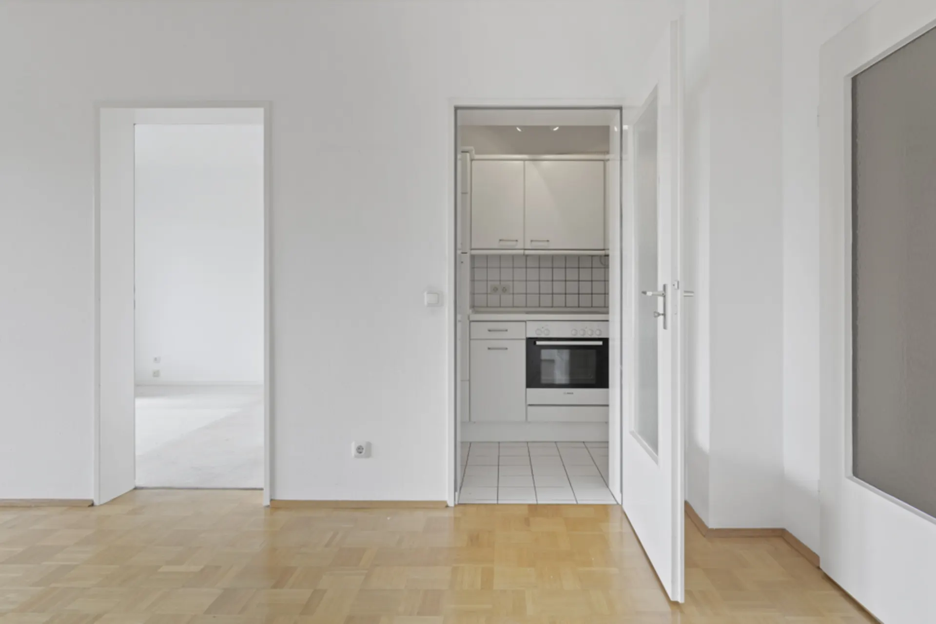 Helles Zimmer mit Parkettboden, offener Tür zur weißen Einbauküche.