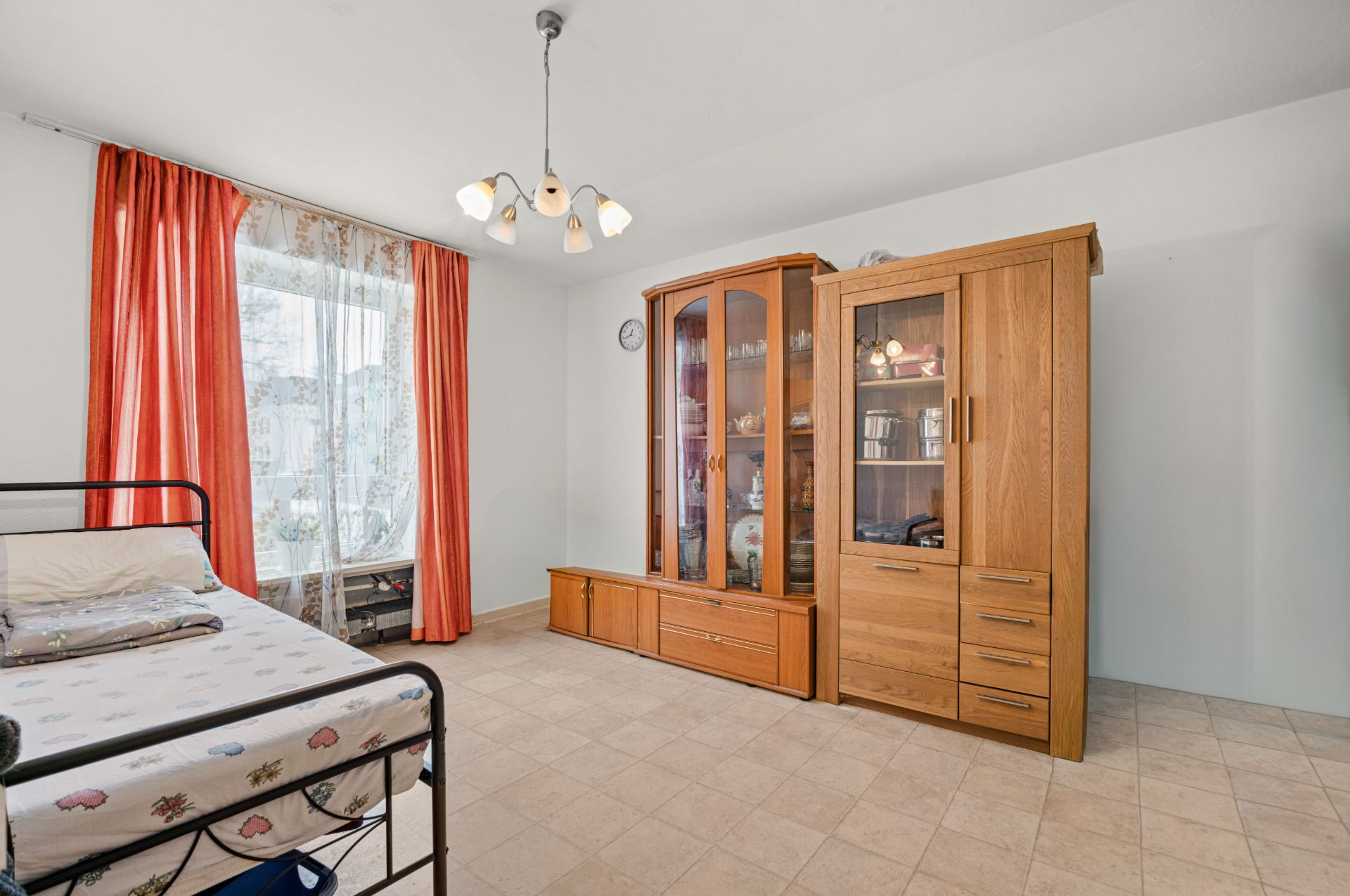 Wohnzimmer mit Einzelbett, großer Holzschrank und Fenster mit roten Vorhängen.