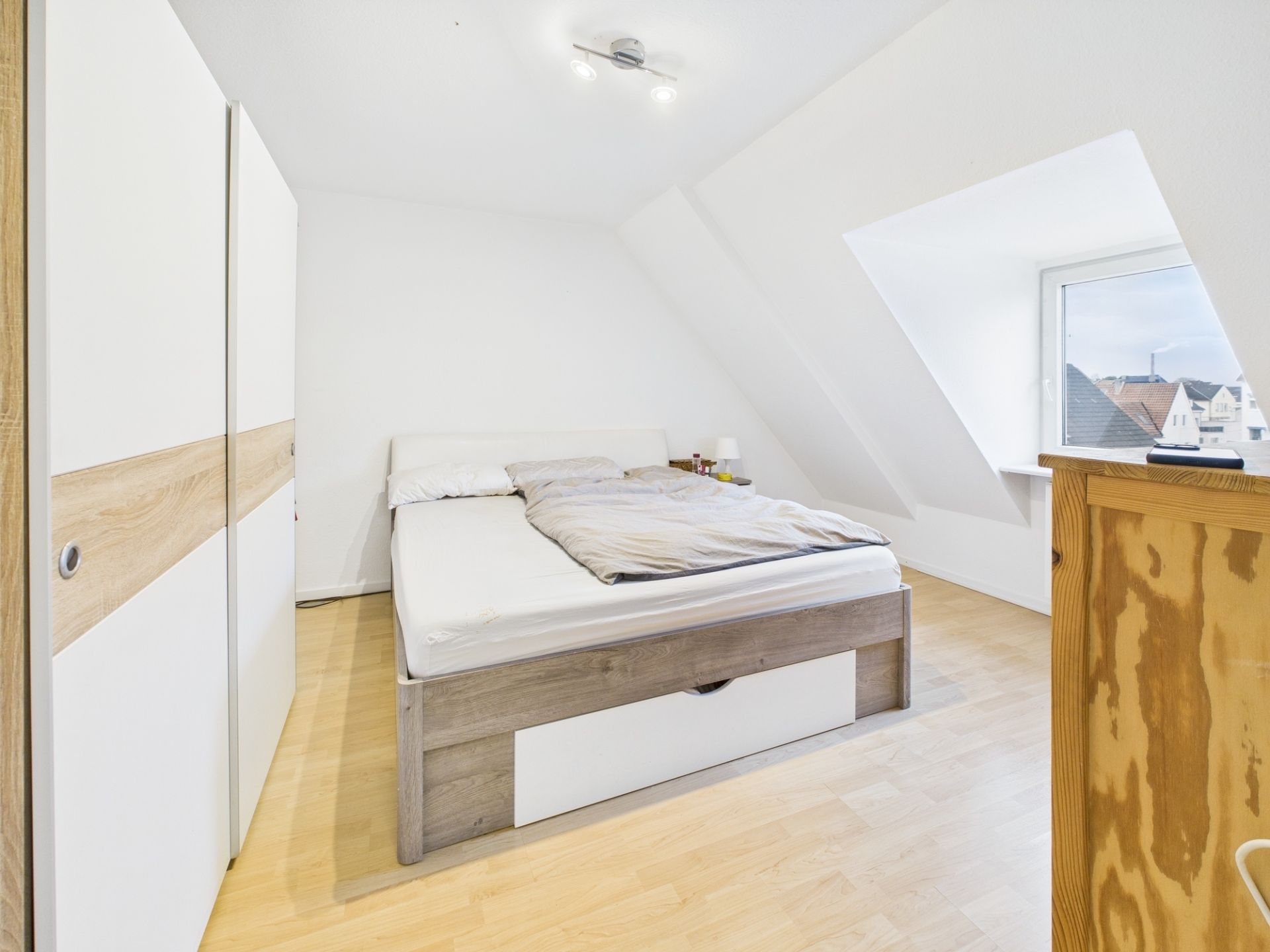 Helles Schlafzimmer mit Bett, Kleiderschrank, Kommode und Fenster im Dachgeschoss.