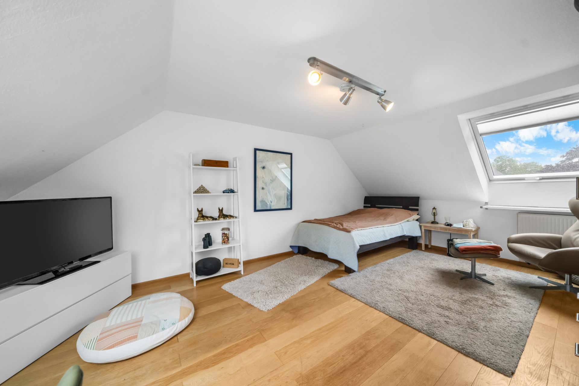 Helles Schlafzimmer mit Dachschräge, Bett, Fernseher, Regal und Sessel auf Holzfußboden.