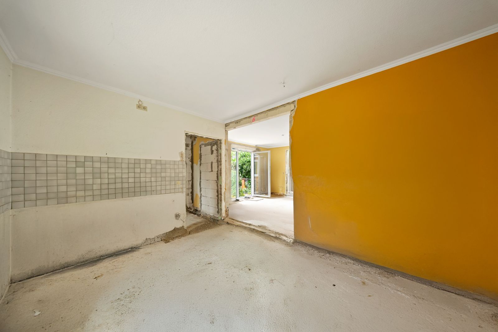 Leerer Raum mit gelber Wand, abgenutztem Boden und offener Tür zum nächsten Zimmer.