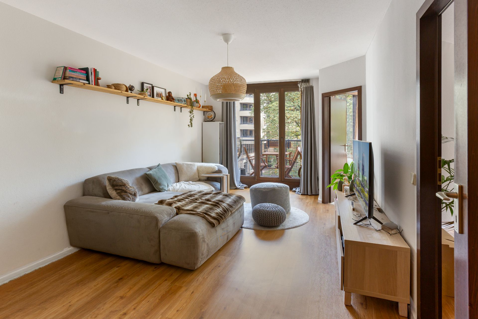 Wohnzimmer mit grauem Sofa, TV, Holzregal und großem Fenster mit Blick auf den Balkon.